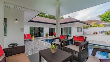 An outdoor pool area with wicker furniture and red cushions, a covered patio, and a house in the background.