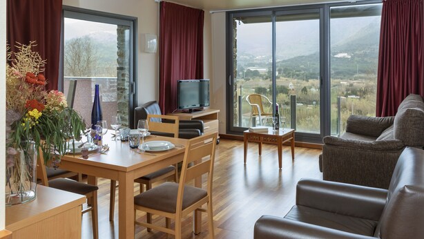 A dining area with a wooden table, chairs, and a television. There is a view of a landscape through large windows.