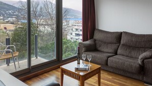 A living room with a sofa, a wooden coffee table, and a view of buildings and mountains through large windows.