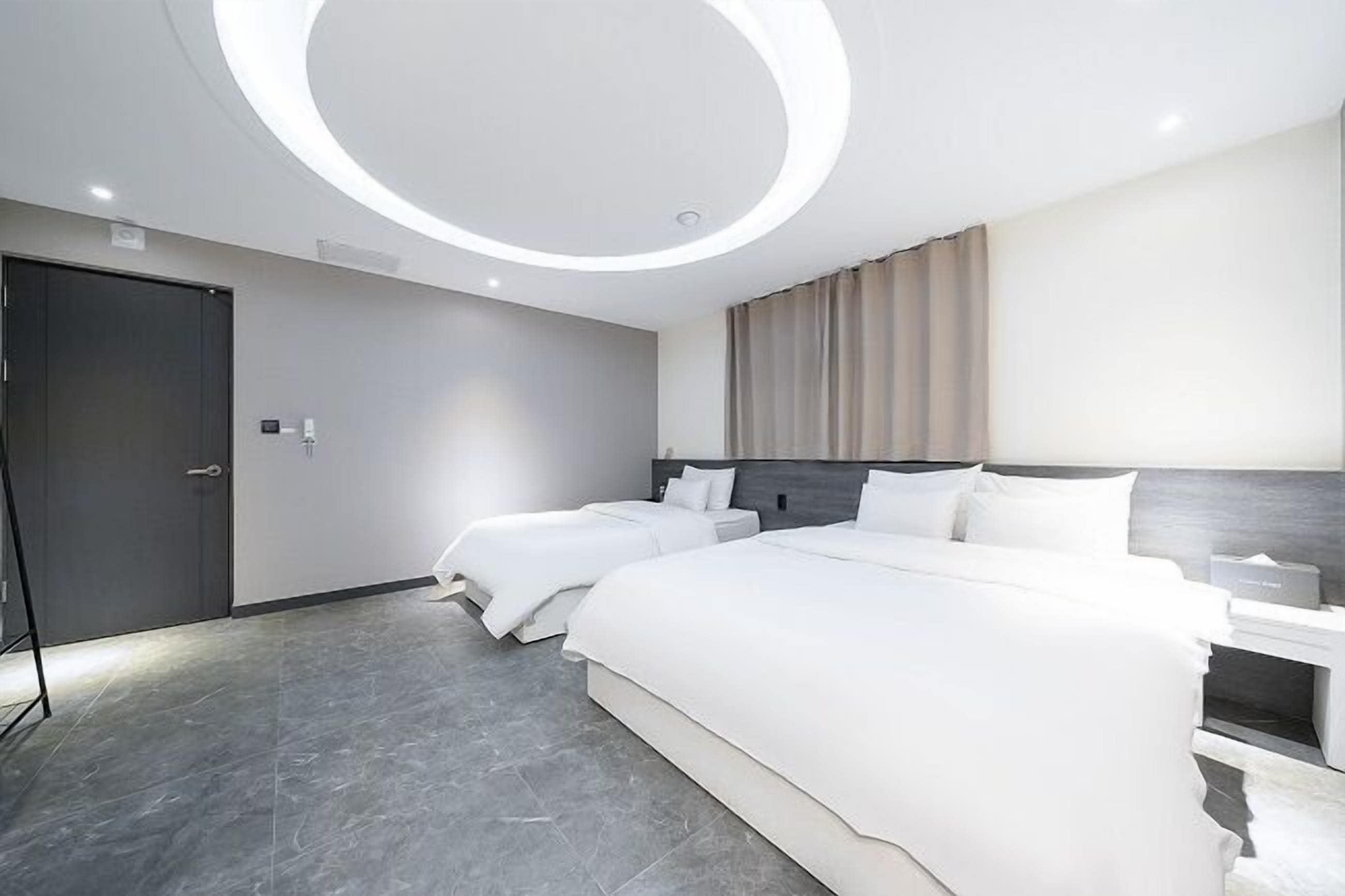 A modern hotel room with a large bed, a smaller bed, a bedside table, a wardrobe, and a circular ceiling light.