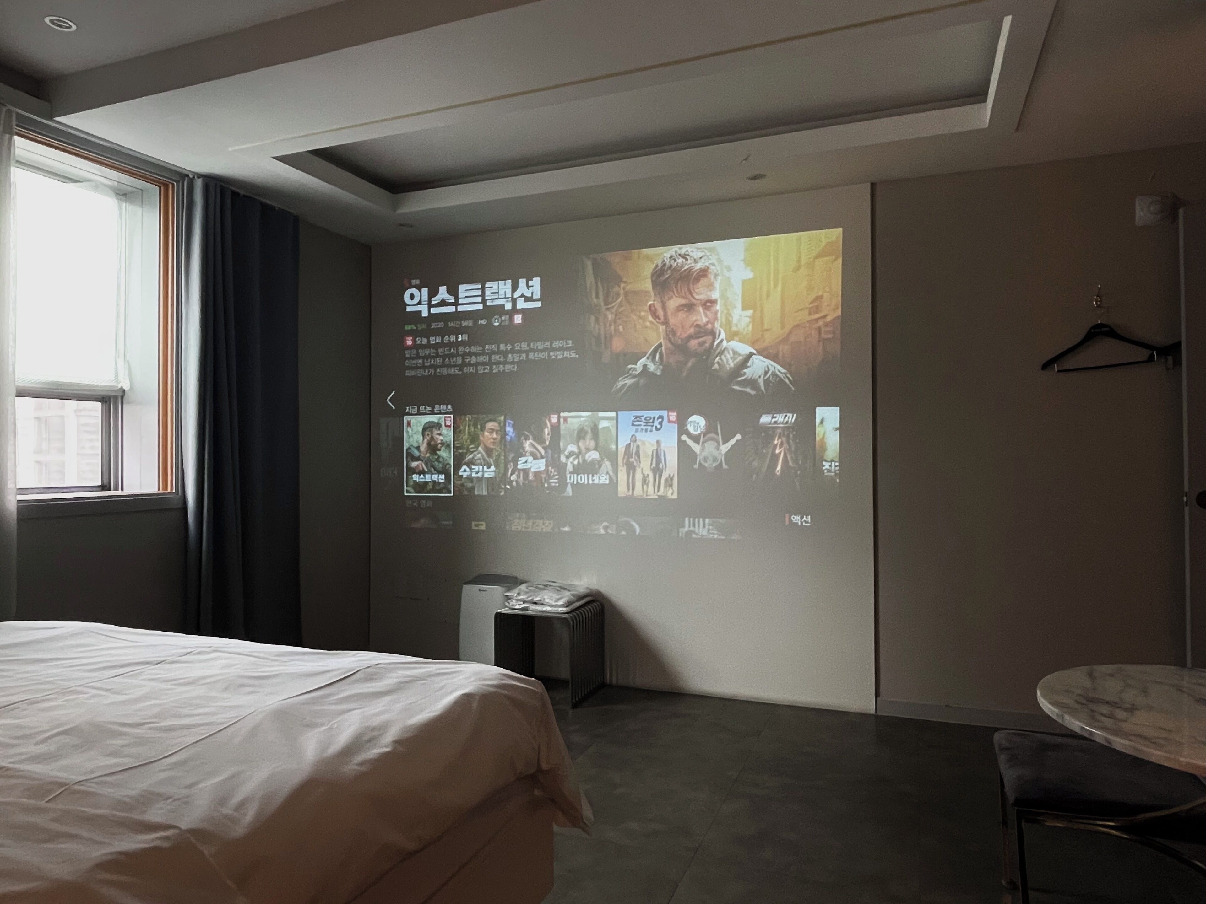 Una habitación de hotel con una cama, una pantalla para proyector mostrando carteles de películas, una mesita y una silla.