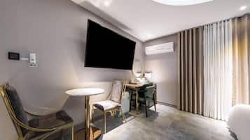 A modern hotel room with a flat-screen TV mounted on the wall, a desk with a chair, a round table, and a large window with curtains.