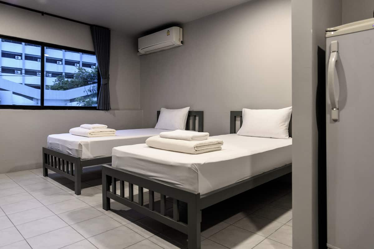 A hotel room with two single beds, a window with a view of buildings, an air conditioning unit, and a refrigerator.