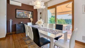 Un espace repas moderne avec une table en verre, des chaises blanches et une grande fenêtre donnant sur l’extérieur.
