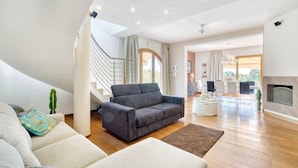 Un vaste espace de vie comprenant un canapé gris, un canapé blanc et un tapis marron. Un escalier mène à un niveau supérieur, et on aperçoit une cheminée au fond.