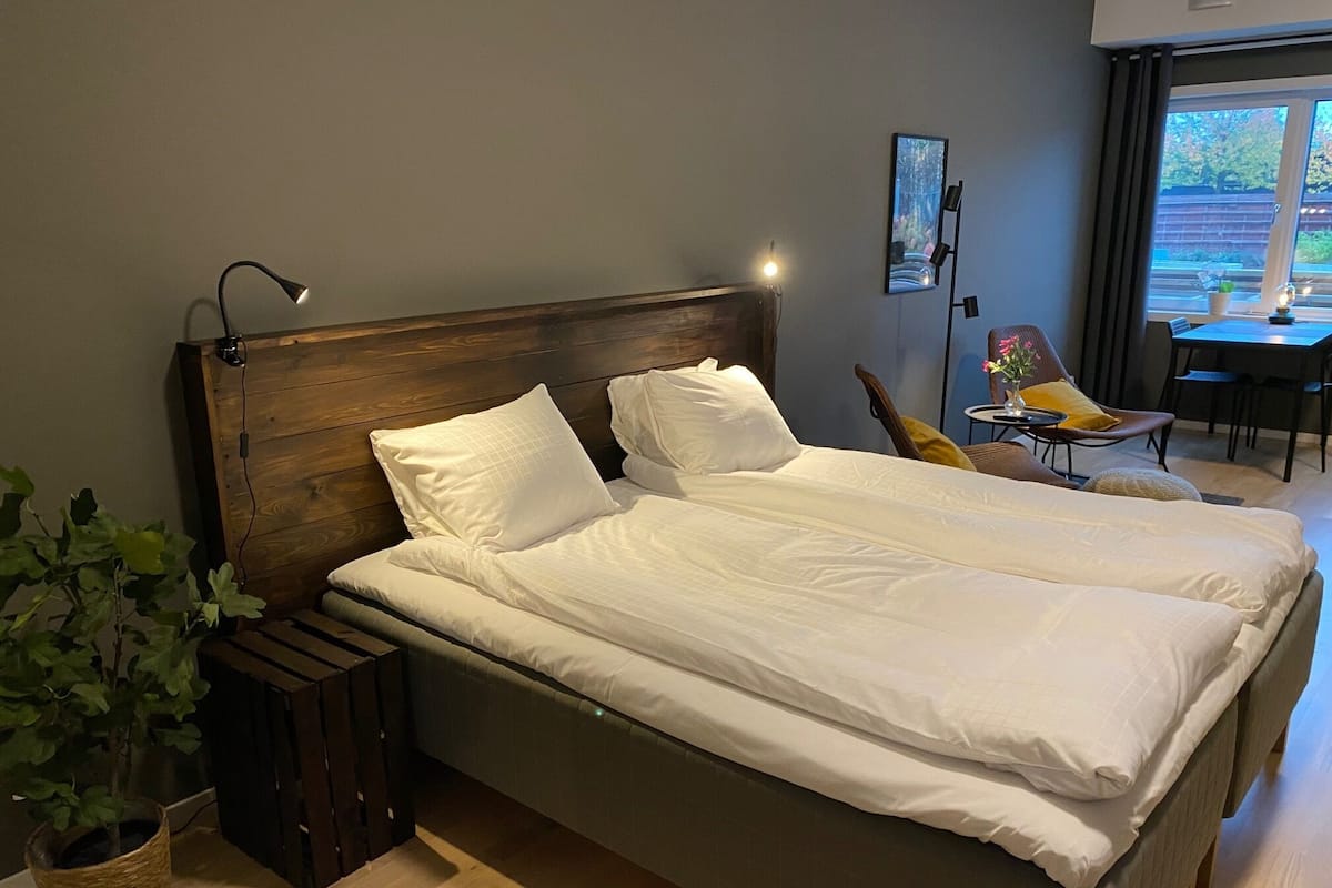 Una cama doble con sábanas blancas, cabecera de madera, lámpara de noche, una mesita con una silla y una planta en maceta.