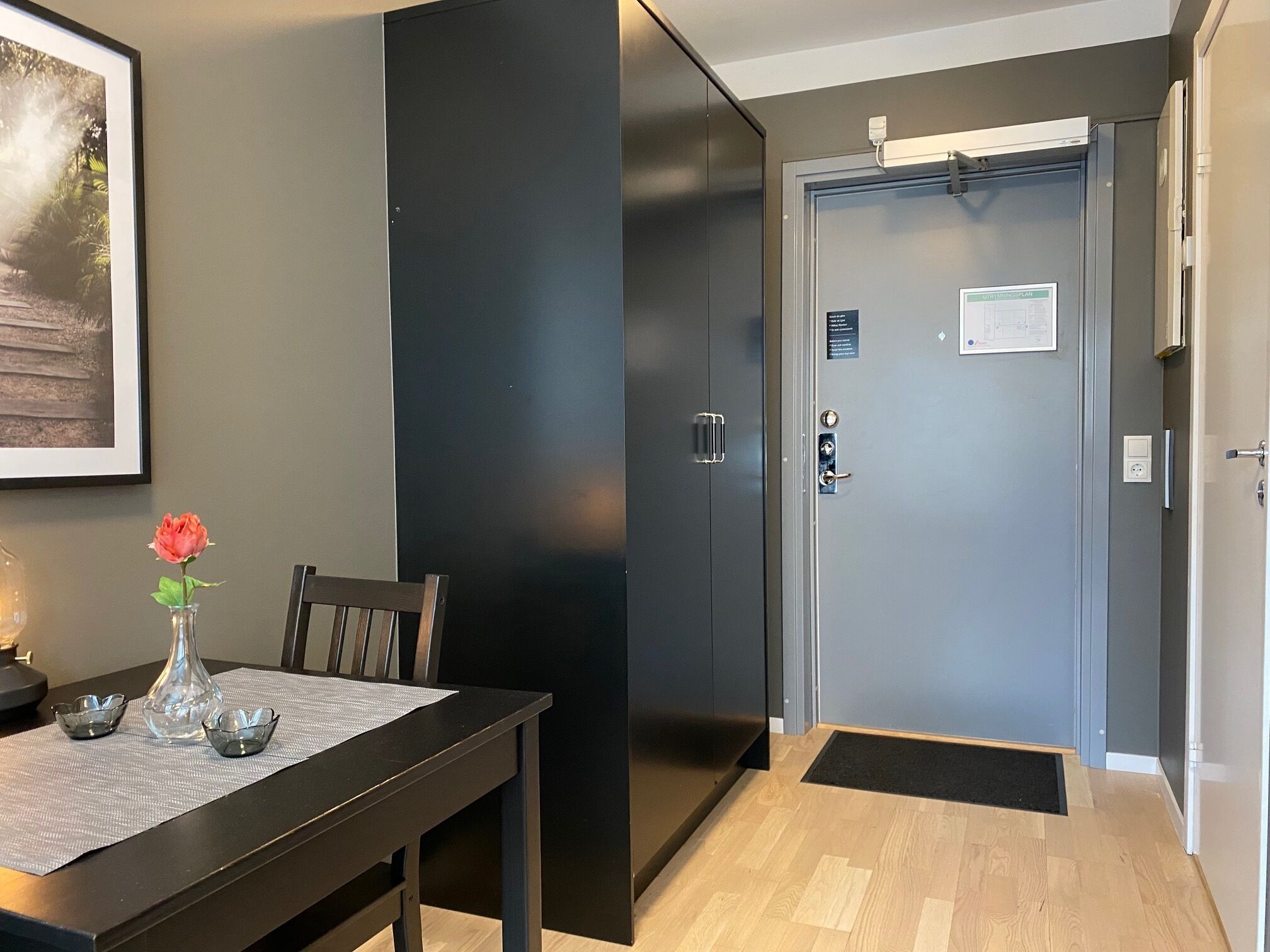 A hotel room with a dark grey door, a small table with a vase of flowers, and a framed picture on the wall.