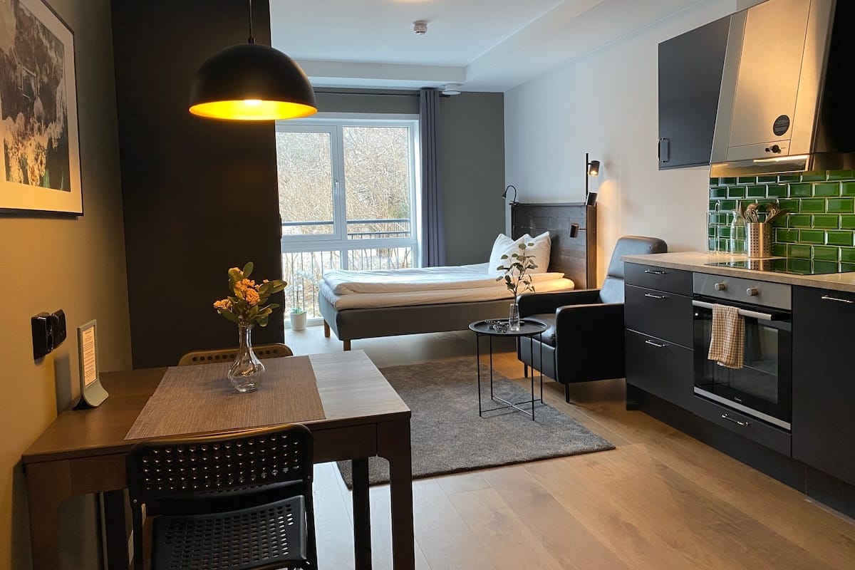 Habitación de hotel compacta con cama, un pequeño comedor, kitchenette y un amplio ventanal con vistas al exterior.