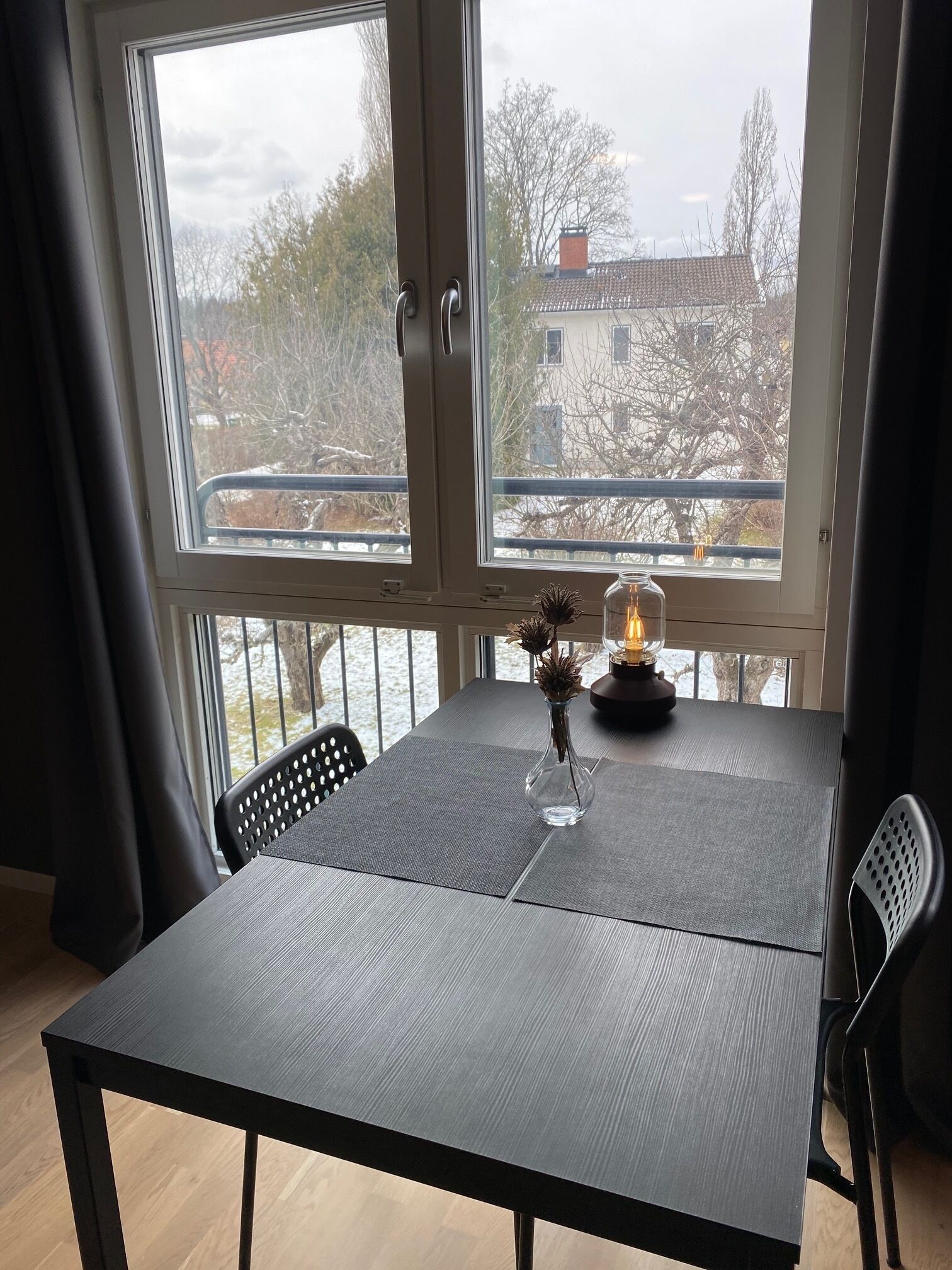 Uma mesa de jantar com um vaso e uma vela, colocada perto de uma janela com vista para árvores e edifícios.