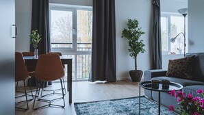 A modern living room with a dining area, large windows with curtains, a potted plant, a sofa with cushions, and a coffee table with remote controls. - Magnolia House Norrviken (Sollentuna)