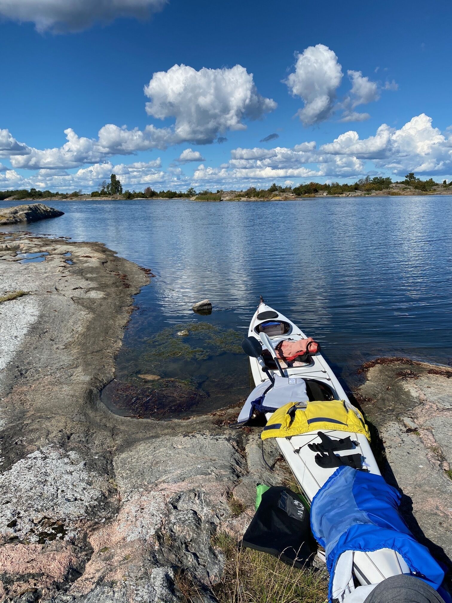 Un kayak reposa sobre una orilla rocosa, con vista despejada a un lago y árboles lejanos bajo un cielo parcialmente nublado.