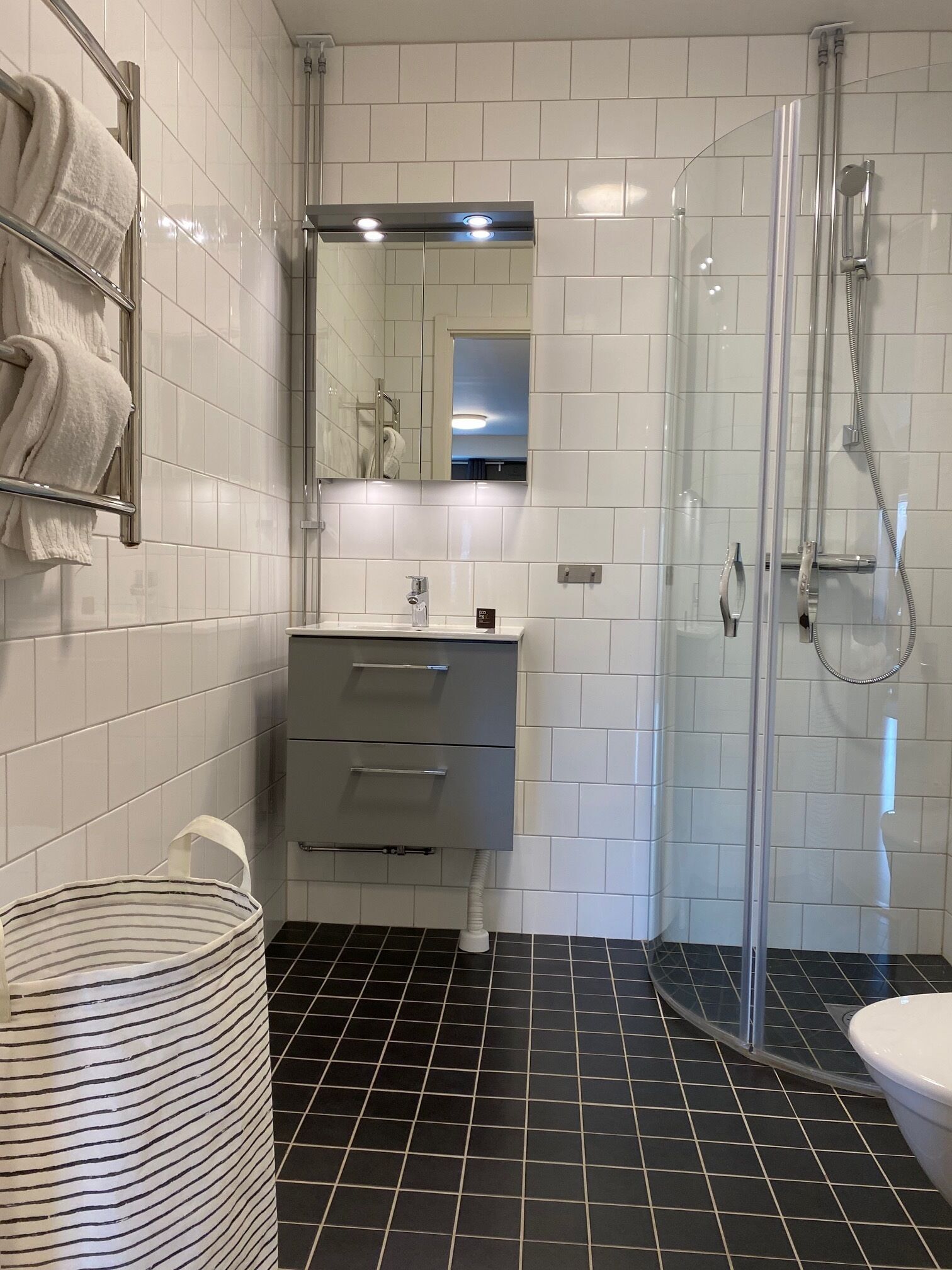 Ein modernes Badezimmer mit weißen Fliesenwänden, einem schwarzen Fliesenboden, einer Duschkabine aus Glas, einem weißen Waschbecken und einem Handtuchhalter mit Handtüchern.