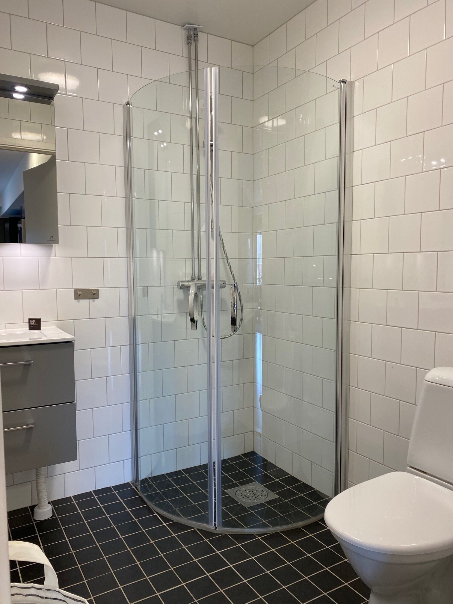 Une salle de bain dotée d’une douche à parois de verre, de murs recouverts de carreaux blancs, d’un sol carrelé noir, de toilettes blanches et d’un meuble gris avec des tiroirs.