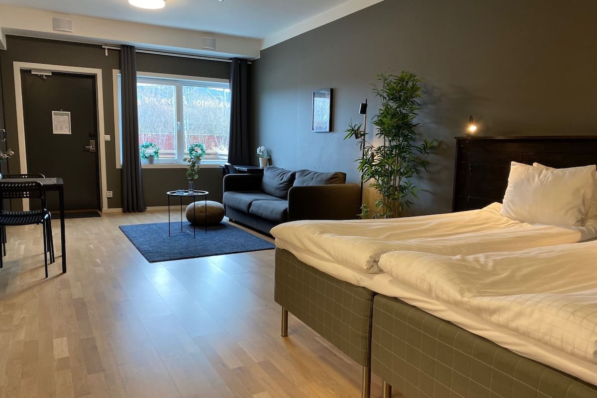 Una habitación de hotel moderna con una cama grande, un sofá, una mesita con una planta y vistas al exterior a través de un ventanal.