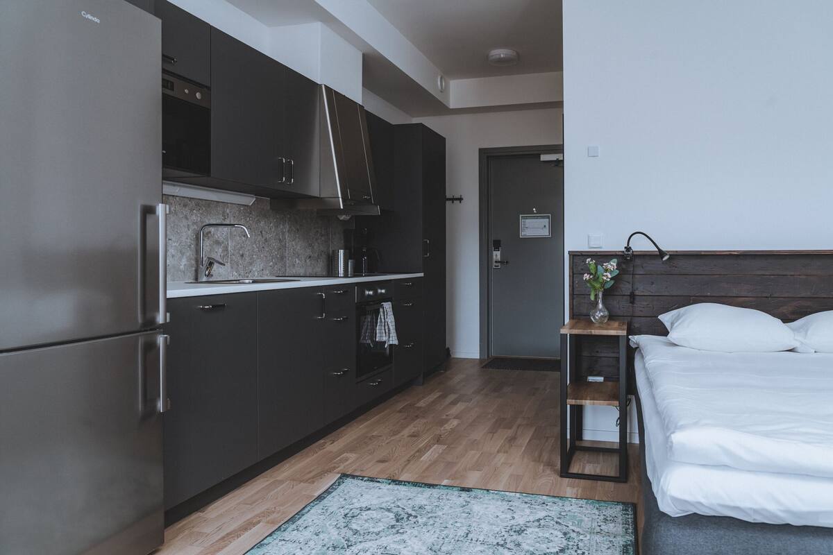 Habitación de hotel pequeña con kitchenette, una cama, mesita de noche con un florero y suelo de madera.