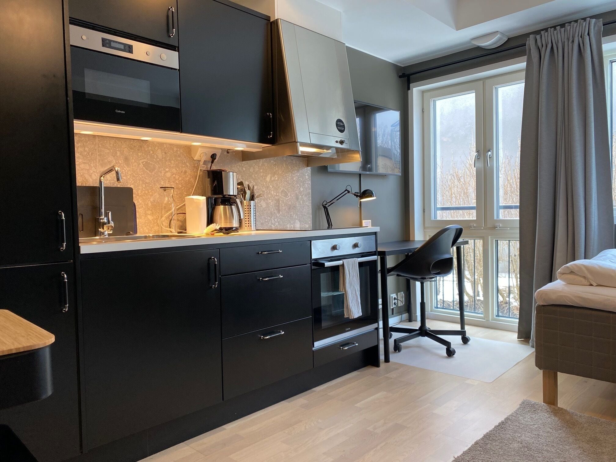 A modern kitchen with black cabinets, a built-in oven, and a coffee maker. There is a window with curtains and a view of trees outside.