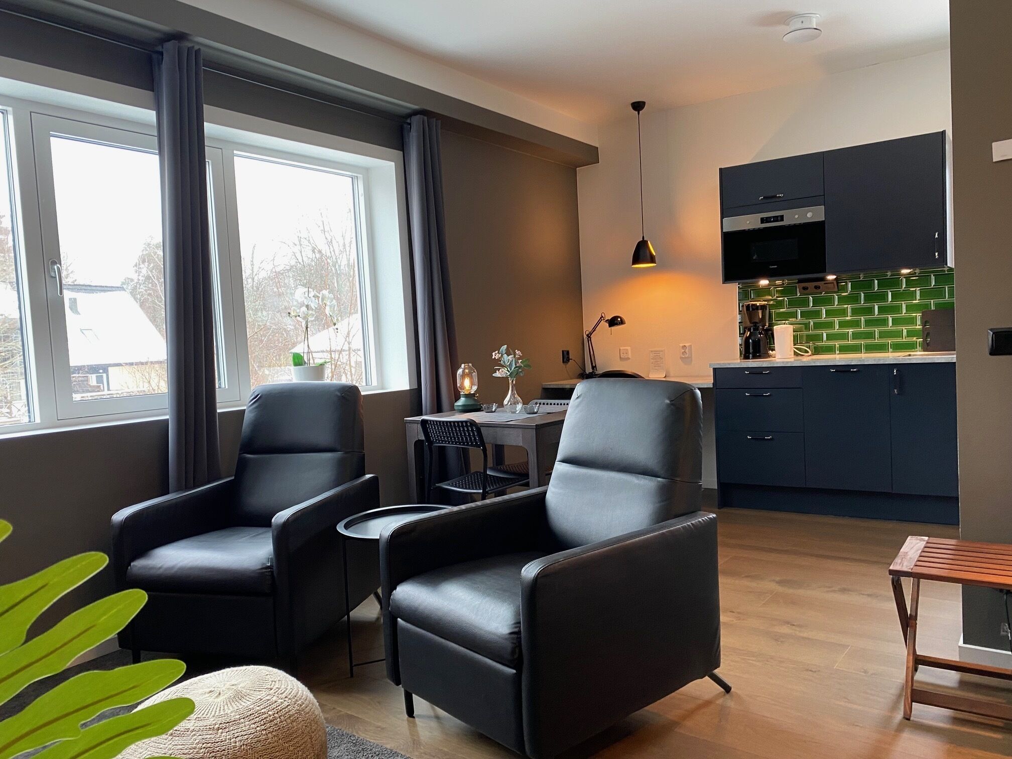 Uma sala de estar moderna com duas poltronas de couro preto, uma mesa de centro de madeira e uma mesa de jantar com cadeiras. Uma cozinha com armários escuros e um revestimento verde é visível ao fundo.