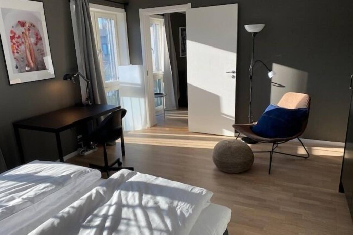Un dormitorio con cama, escritorio, silla, un otoman redondo y una puerta que da a otra habitación.