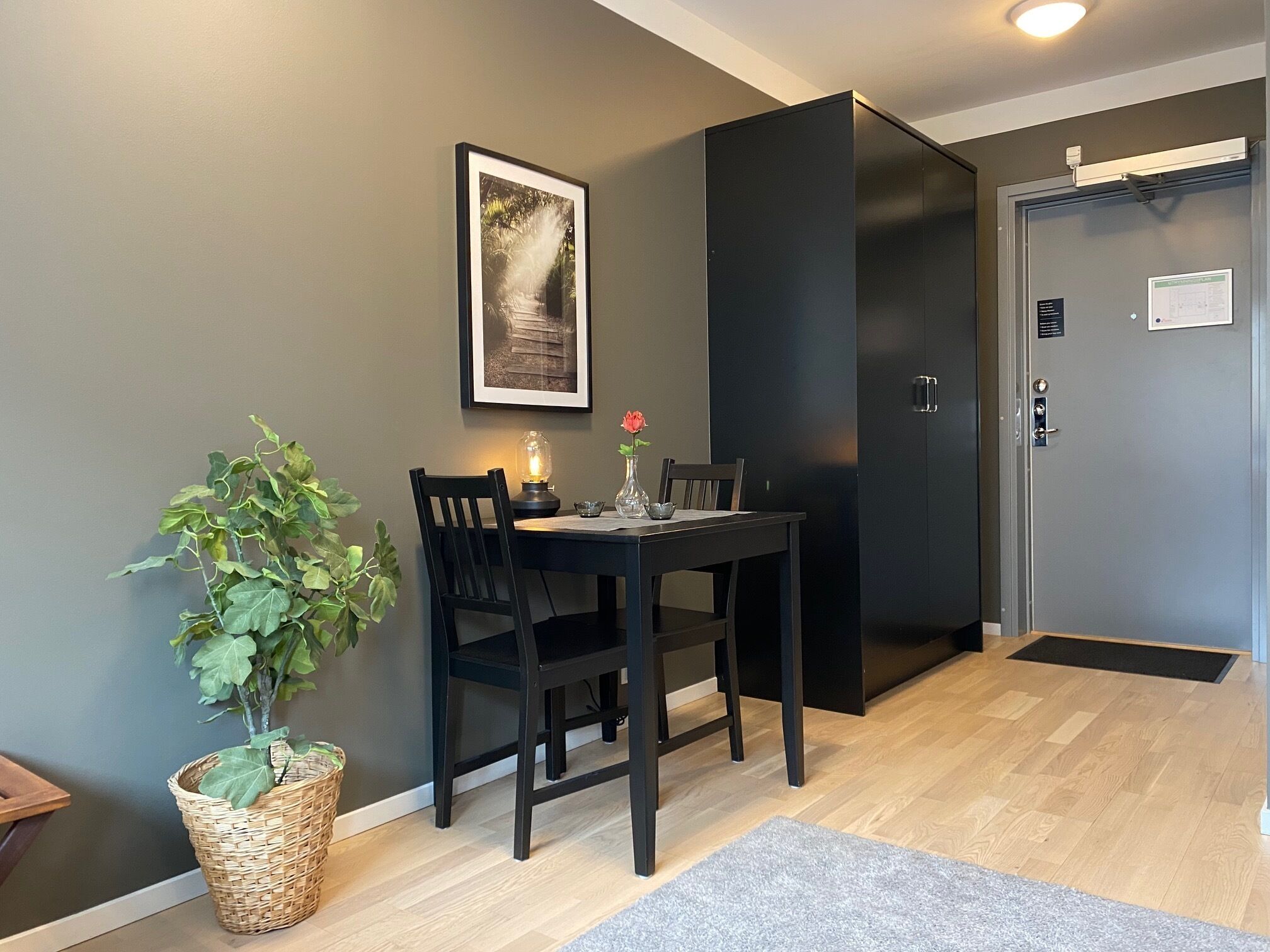 Comedor con una mesa, sillas, planta en maceta, un cuadro enmarcado y una puerta con cámara de seguridad.