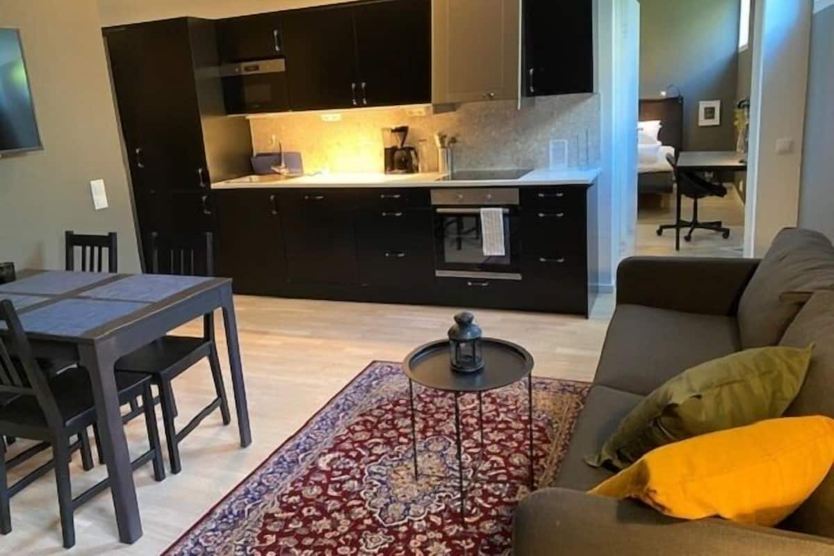 Una cocina moderna con armarios oscuros y una isla central, un comedor con mesa y sillas, y una sala de estar con sofá y alfombra estampada.