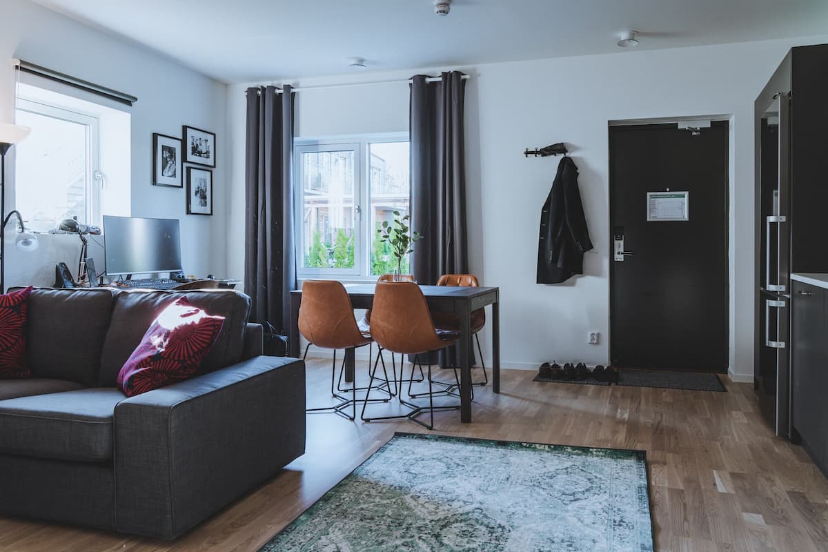 Una sala de estar moderna con un sofá gris, una mesa de comedor con dos sillas, un ventanal con cortinas y una puerta negra con un abrigo colgado.
