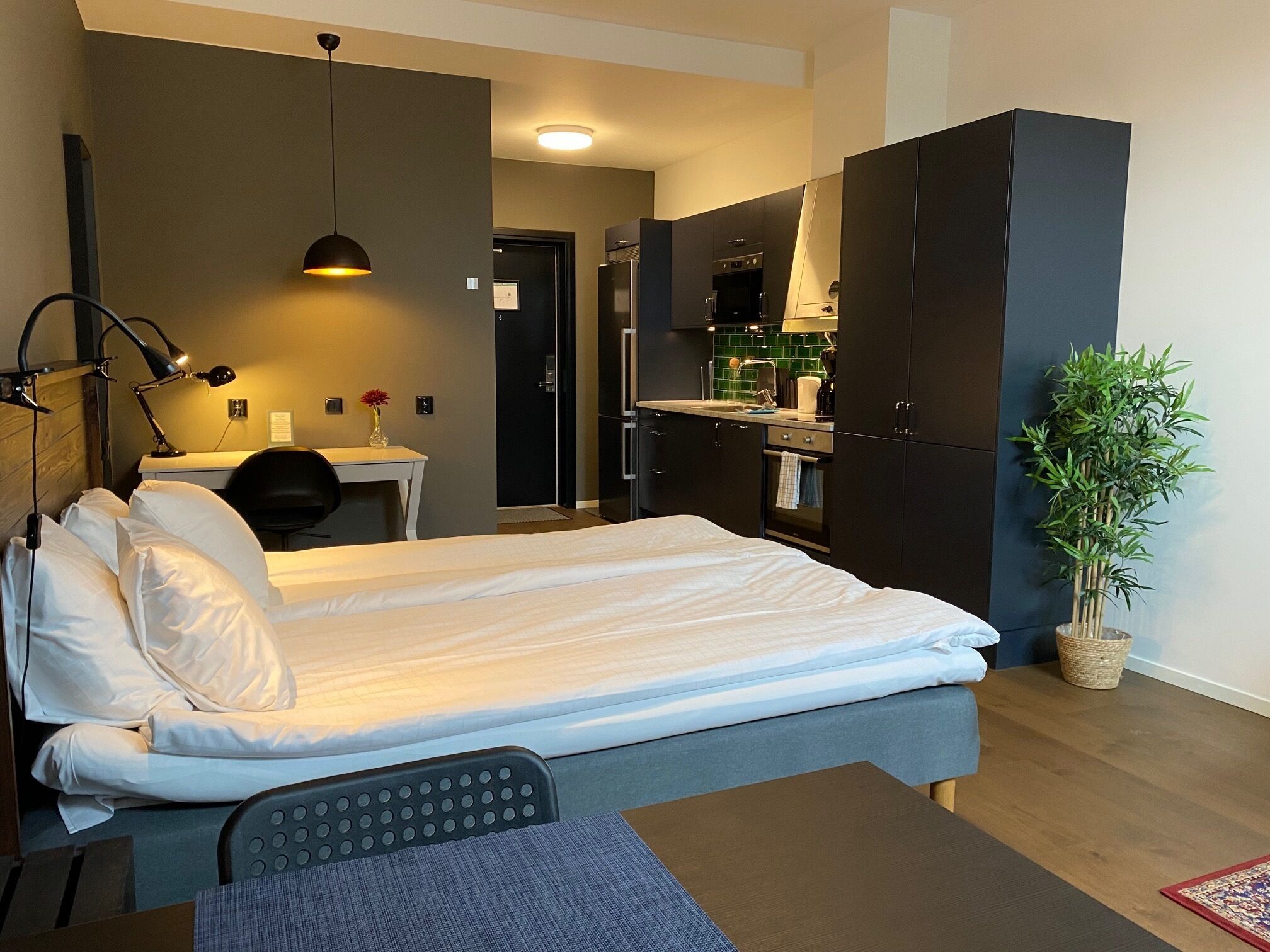 A modern hotel room with a bed, desk, and kitchenette.