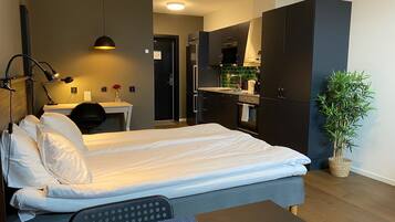 A modern hotel room with a bed, desk, and kitchenette.