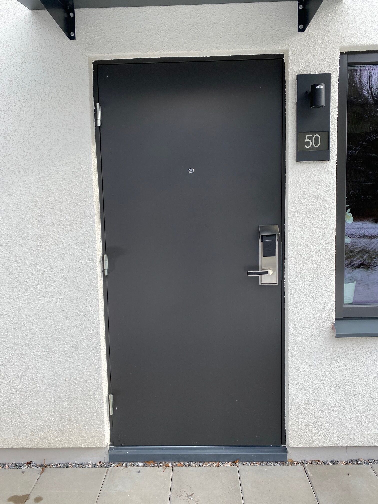 A modern black door with a numeric keypad lock, a small window above it, and a number plate '50' on the wall.