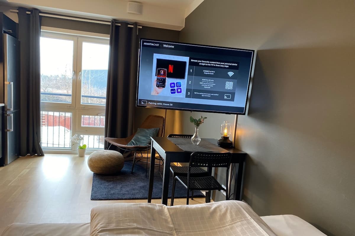Una habitación de hotel con una cama, un escritorio con computadora, una silla, un televisor montado en la pared y una ventana con cortinas.