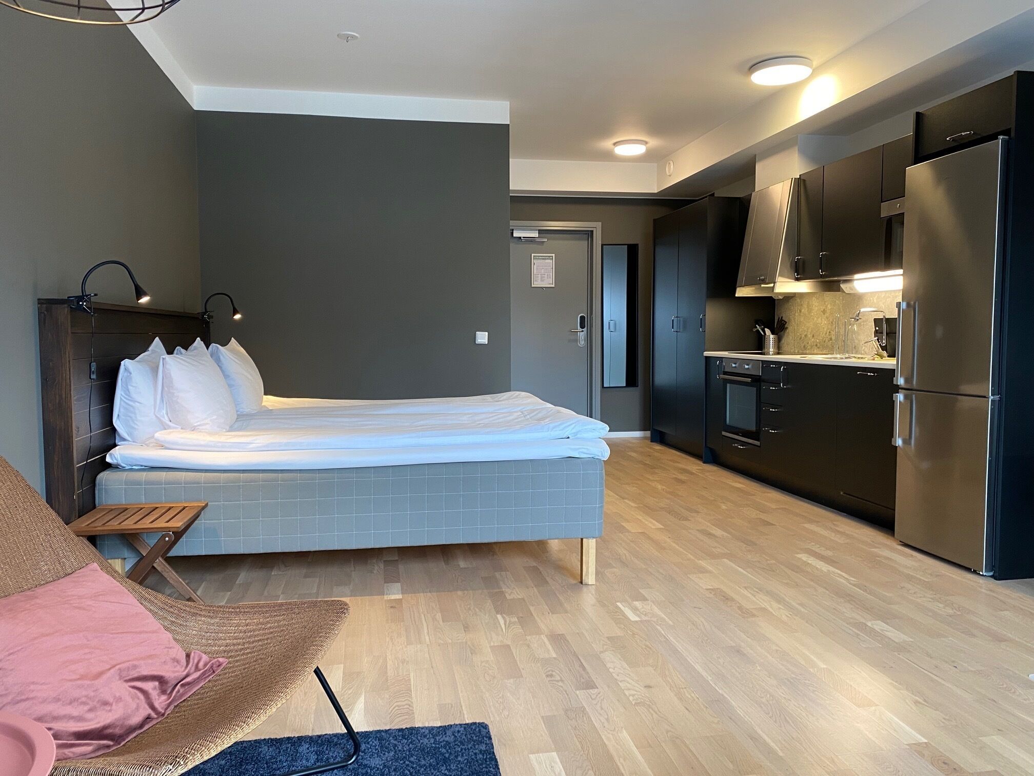 A modern hotel room with a bed, a small seating area, a kitchenette, and a bathroom.