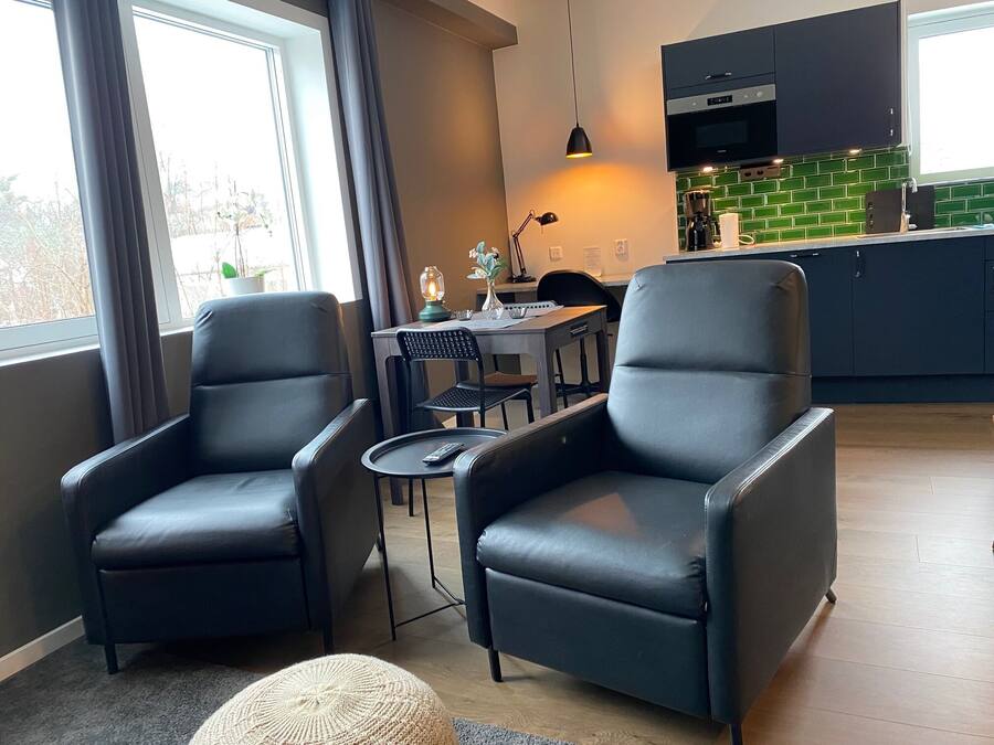 Una sala moderna con dos sillones de cuero negro, una mesita redonda y un escritorio con una lámpara. Se observa una cocina al fondo, con armarios oscuros y un salpicadero verde.