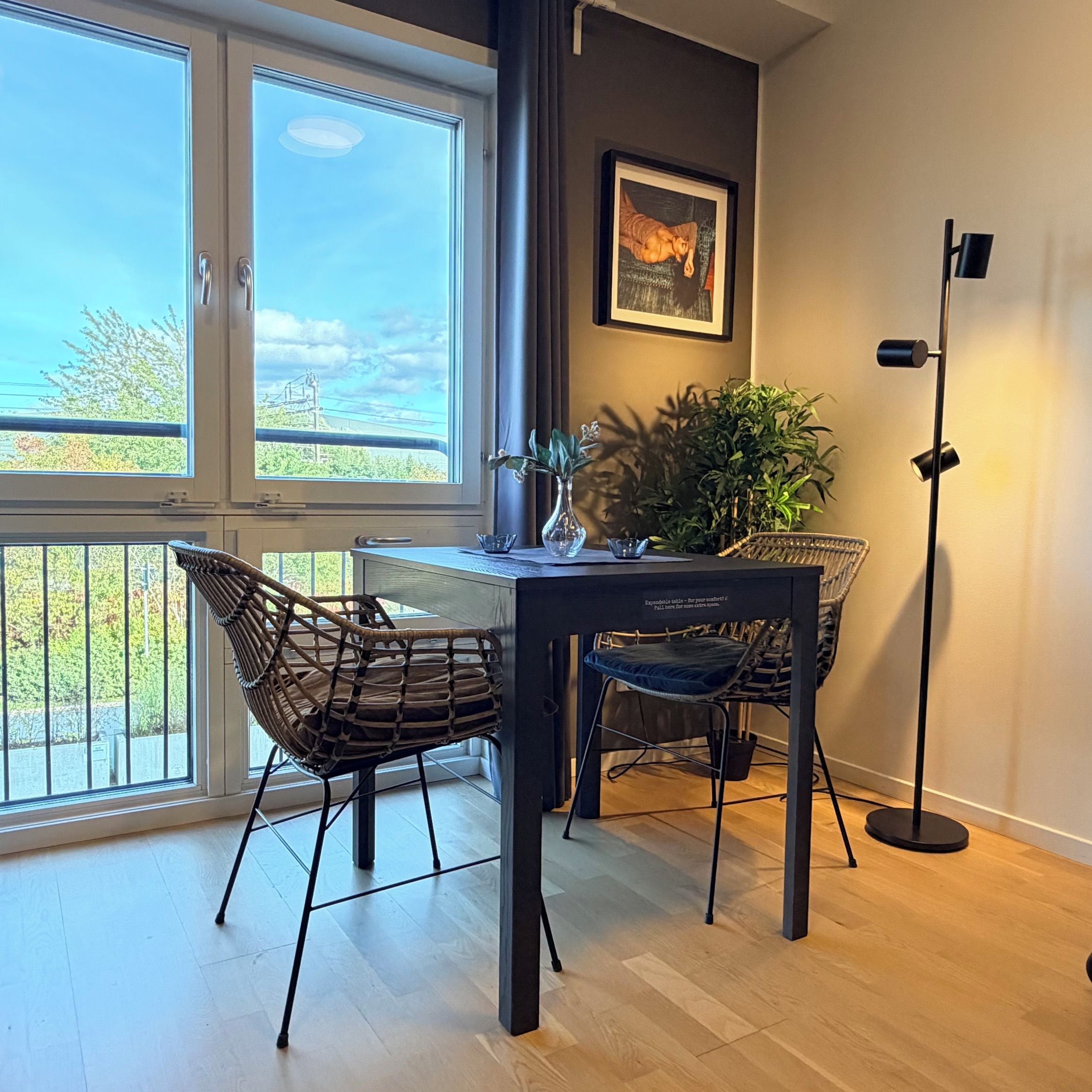Uma área de refeições com vista para o exterior, uma mesa com cadeiras, um abajur e um quadro na parede.