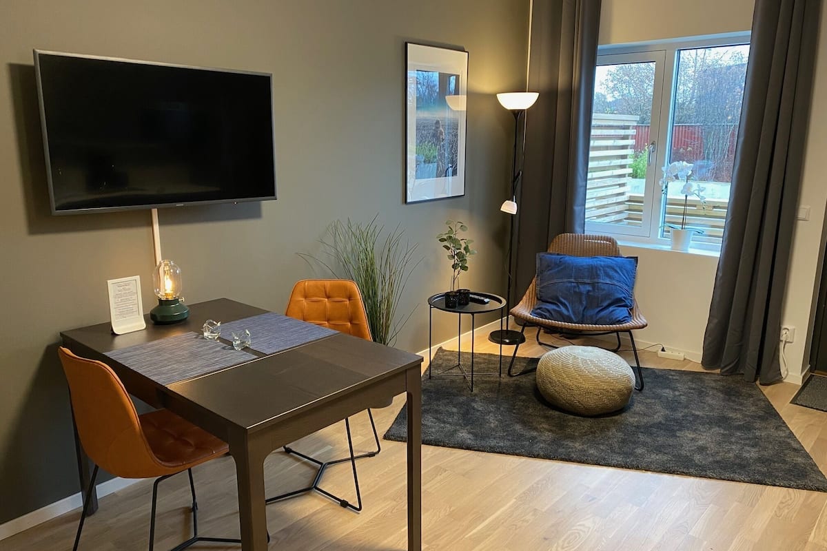 Una sala de estar moderna con un televisor de pantalla plana, una mesa de comedor con sillas, un sofá, una lámpara y un gran ventanal con cortinas.