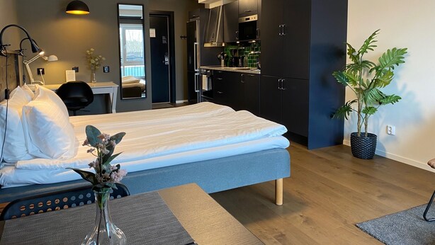 A modern hotel room with a bed, desk, chair, and kitchenette. - Magnolia House Norrviken (Sollentuna)