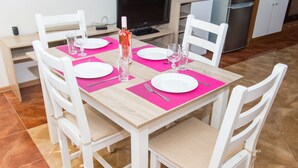 Ein Esstisch ist mit weißen Tellern, rosa Untersetzern und klaren Gläsern gedeckt, umgeben von weißen Stühlen mit gepolsterten Sitzflächen.