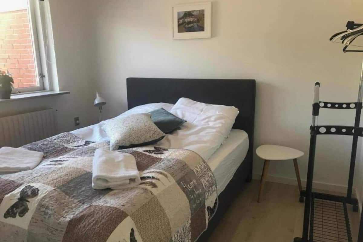 A neatly made bed with a quilt, pillows, and folded towels. A small round table and a black metal rack with clothes are visible.