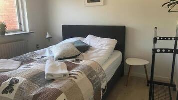 A neatly made bed with a quilt, pillows, and folded towels. A small round table and a black metal rack with clothes are visible.