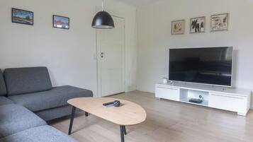 Un salón con un sofá gris, una mesa de centro de madera, un televisor de pantalla plana sobre un soporte blanco y una puerta con marco blanco.