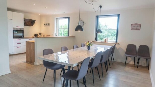 Une cuisine moderne dotée d’un four encastré, un coin repas avec une grande table et des chaises, et de grandes fenêtres laissant entrer la lumière naturelle.