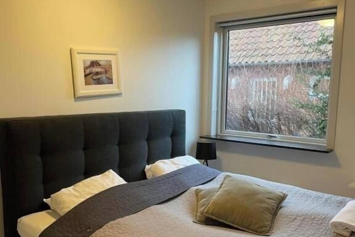A neatly made bed with a dark headboard, a window with a view of a building, and a framed picture on the wall.