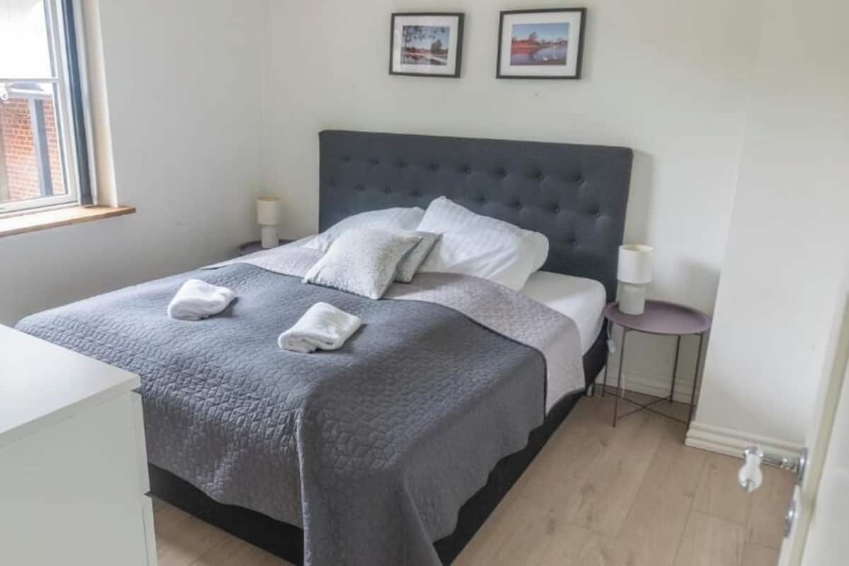 A neatly made bed with a grey quilt and two pillows, a bedside table with a lamp, and a small round table with a lamp.