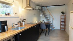 Une cuisine moderne avec un plan de travail en bois, des armoires noires, un lave-vaisselle intégré et un évier en acier inoxydable. On y trouve un coin repas avec une table et des chaises, un escalier et une petite plante posée sur une étagère.