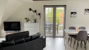Un salon moderne avec un canapé en cuir noir, un téléviseur à écran plat, un coin repas comprenant une table et des chaises blanches, et un balcon offrant une vue sur l’extérieur.