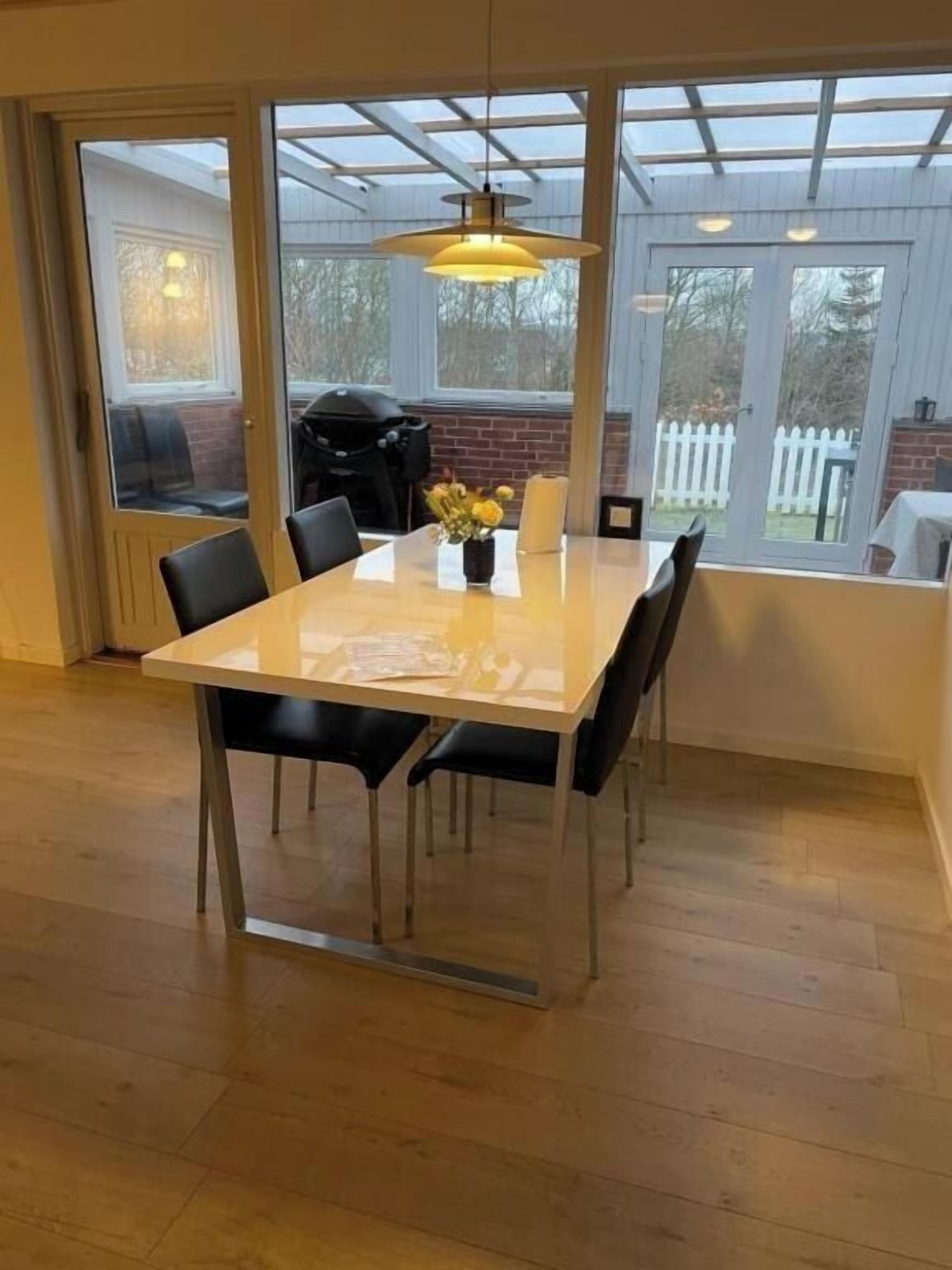 Un comedor con una mesa y sillas, una lámpara colgante y vistas al exterior a través de grandes ventanales.