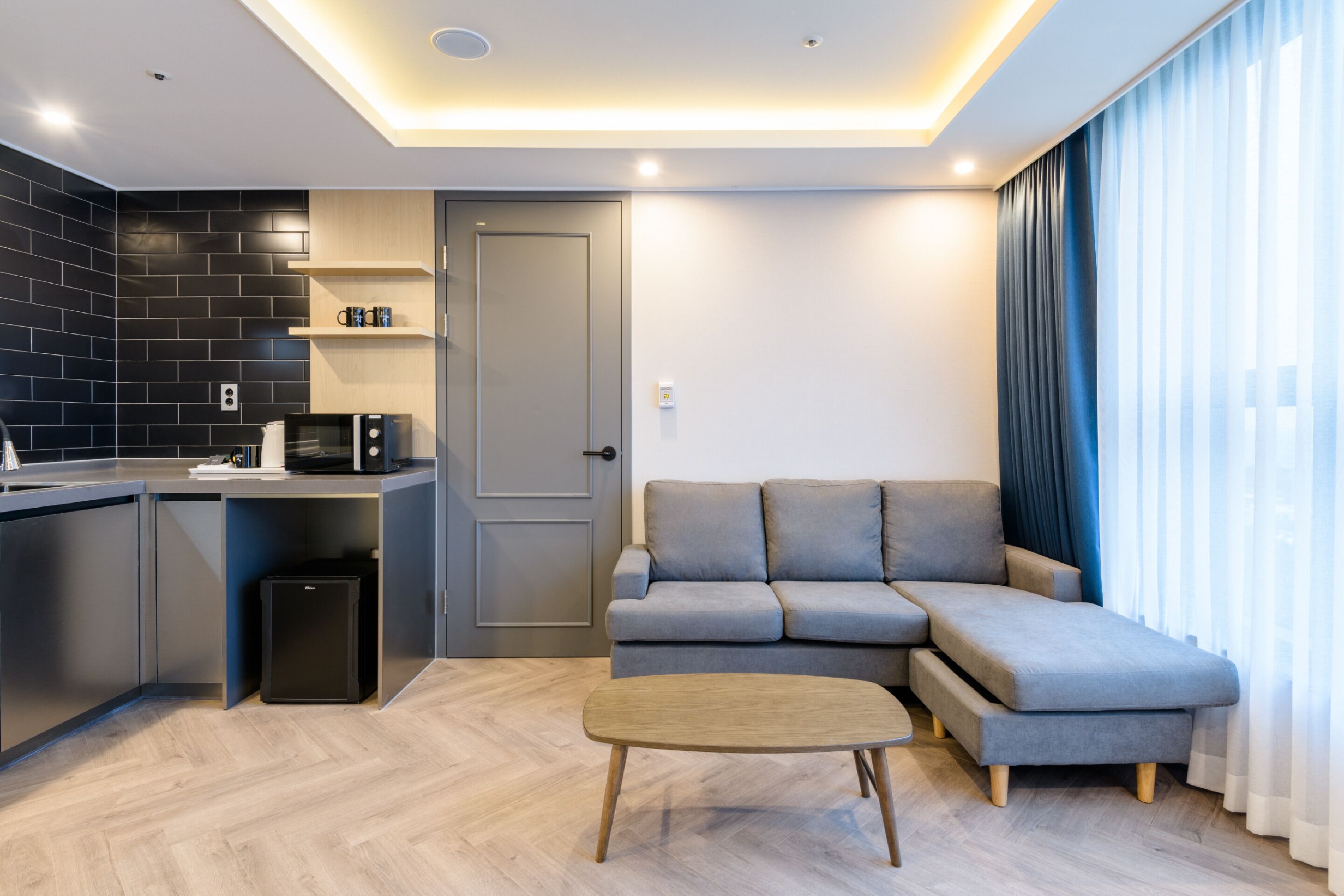 Una sala de estar moderna con un sofá seccional gris, una pequeña mesa de centro de madera, y una zona de cocina con microondas y cafetera.