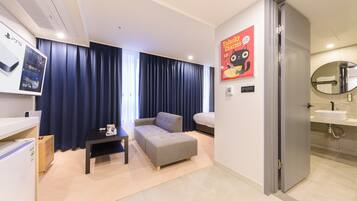 Ein modernes Hotelzimmer mit grauem Sofa, einem kleinen Tisch, einem an der Wand befestigten Fernseher und einem Badezimmer mit Waschbecken und Spiegel.