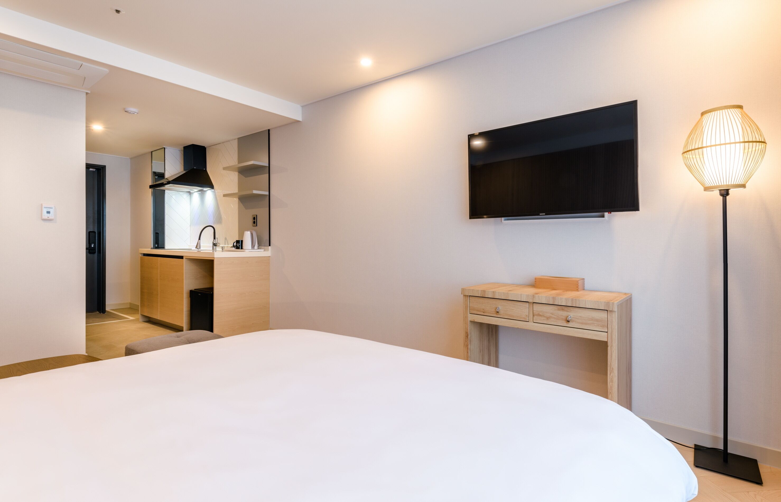 A modern hotel room with a large bed, a wooden nightstand, a flat-screen TV mounted on the wall, and a floor lamp.