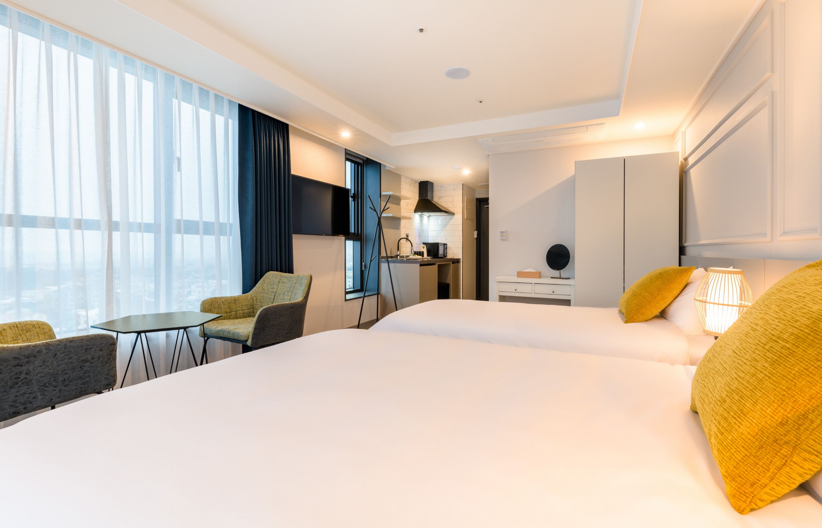A hotel room with two beds, a small table, a chair, a desk, and a kitchenette.