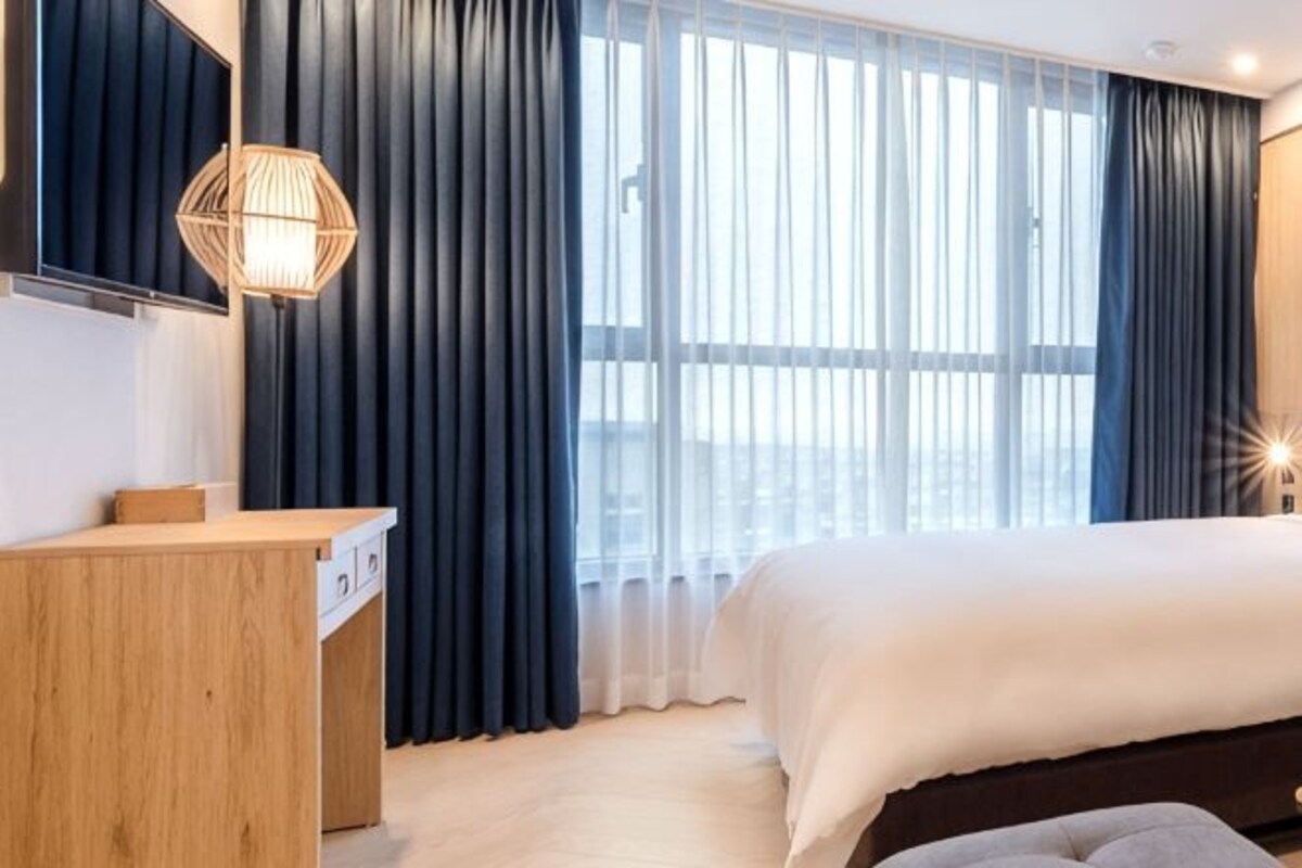Habitación de hotel con cama, escritorio, lámpara y un amplio ventanal con cortinas.