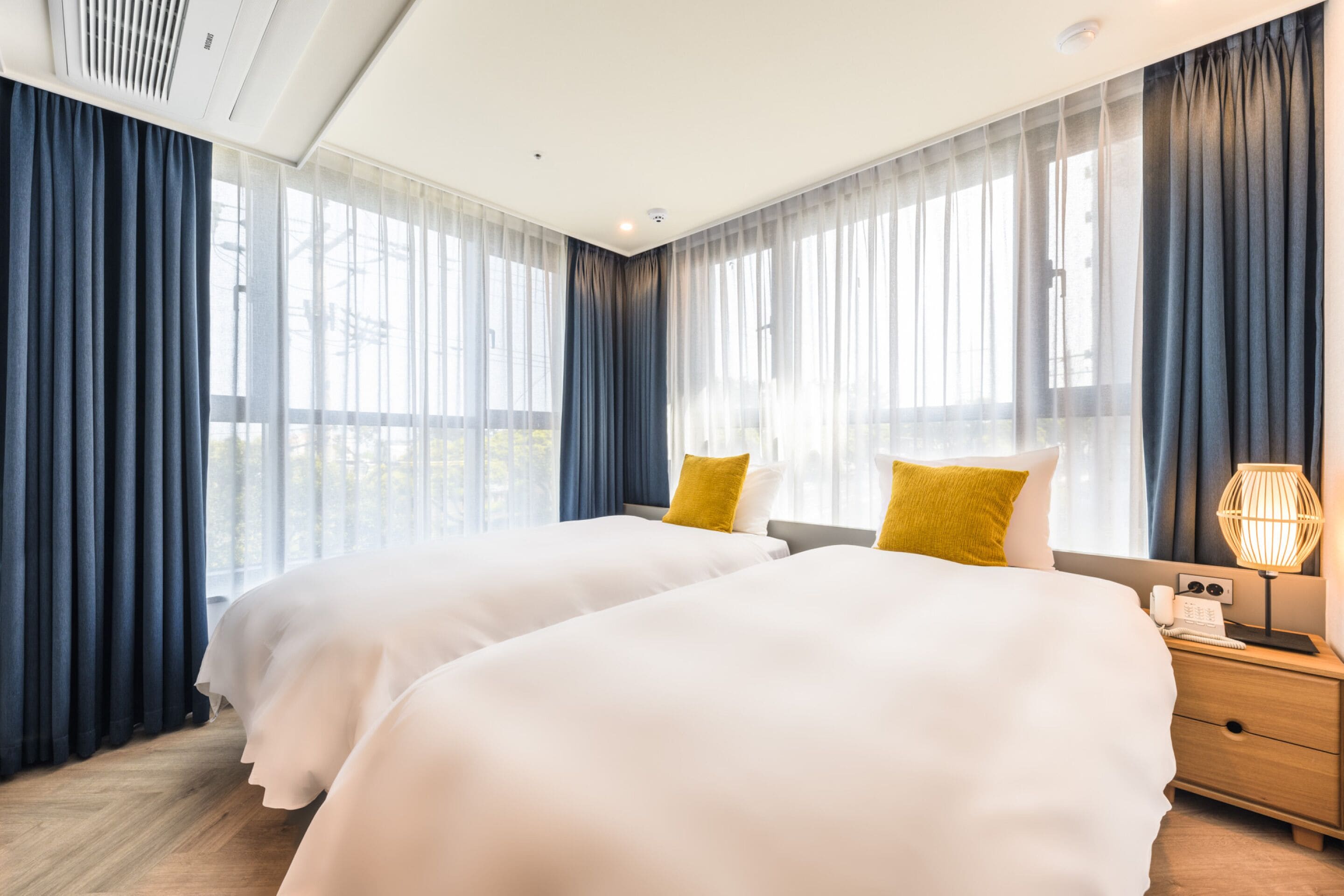 Habitación de hotel con dos camas, una mesita de noche de madera, una lámpara y ventanas grandes con cortinas de gasa.